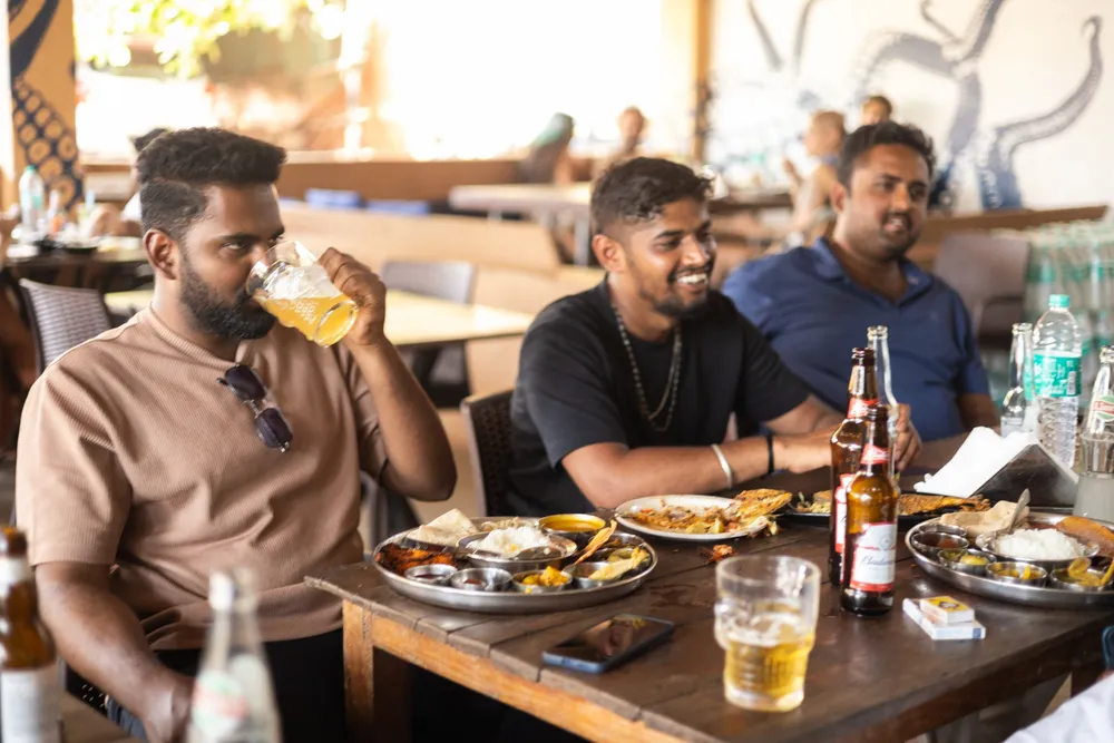 Three men at a wooden table with steel thalis and cold beer.