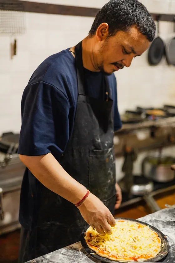 A cook spreading cheese across a pizza base on the kitchen counter.