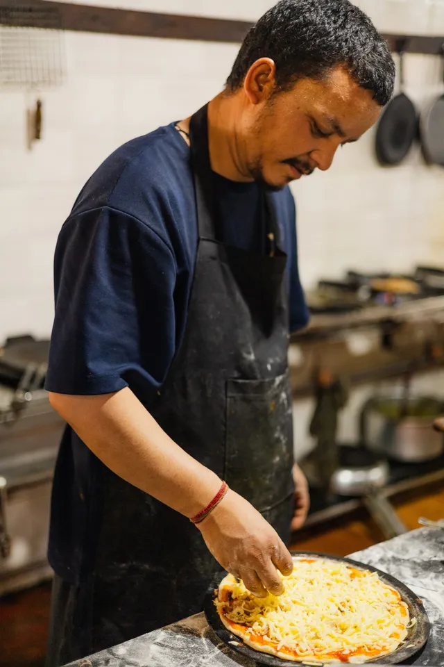A cook spreading cheese across a pizza base on the kitchen counter.
