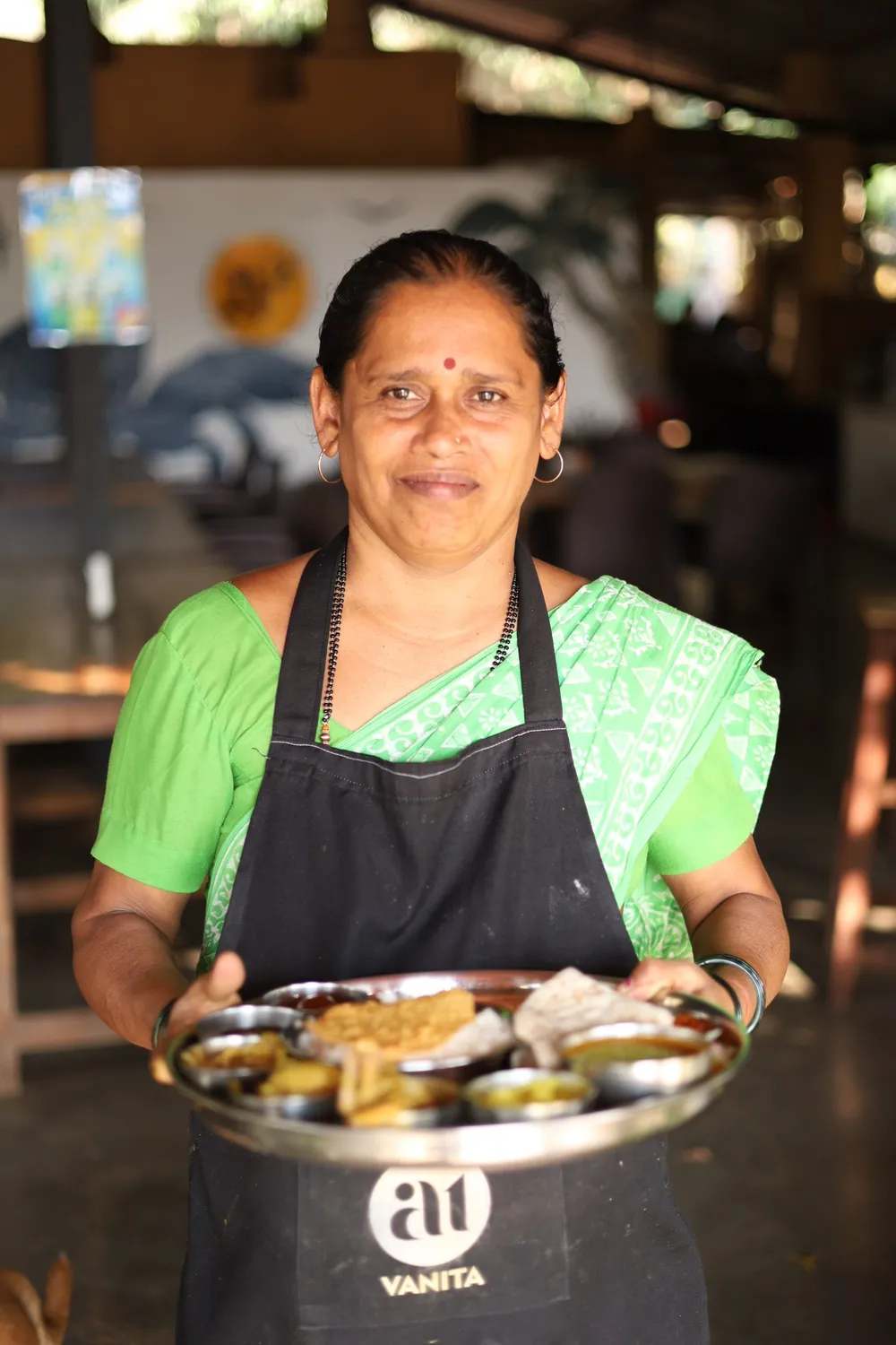 Vanita, in a green sari and an a1 apron, carrying a full steel thali.